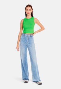Green ribbed crop top, light blue wide-leg jeans with a frayed hem, and patterned sneakers. Simple background, model stands with one hand on hip.