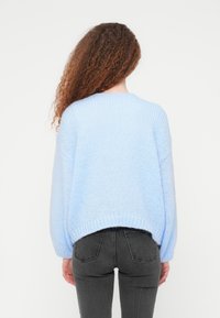 Γυναίκα με μακριά σγουρά μαλλιά, που φοράει ένα ανοιχτό μπλε πλεκτό πουλόβερ και σκούρα γκρι τζιν, στέκεται με την πλάτη προς τα εμπρός σε απλό φόντο.