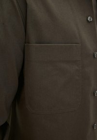 Nærbillede af en mørkebrun jakke med en stor firkantet brystlomme og synlige knapper foran.