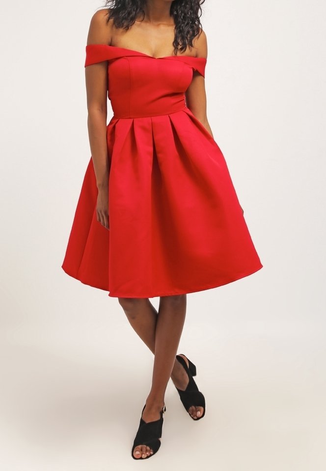 Femme portant une robe rouge mi-longue à épaules dénudées avec des plis, associée à des talons ouverts noirs, debout devant un fond uni.