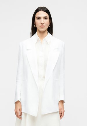 Femme aux cheveux bruns lisses portant des boucles d'oreilles anneaux dorées, une chemise blanche à col et un blazer blanc surdimensionné, regardant vers l'avant sur un fond uni.