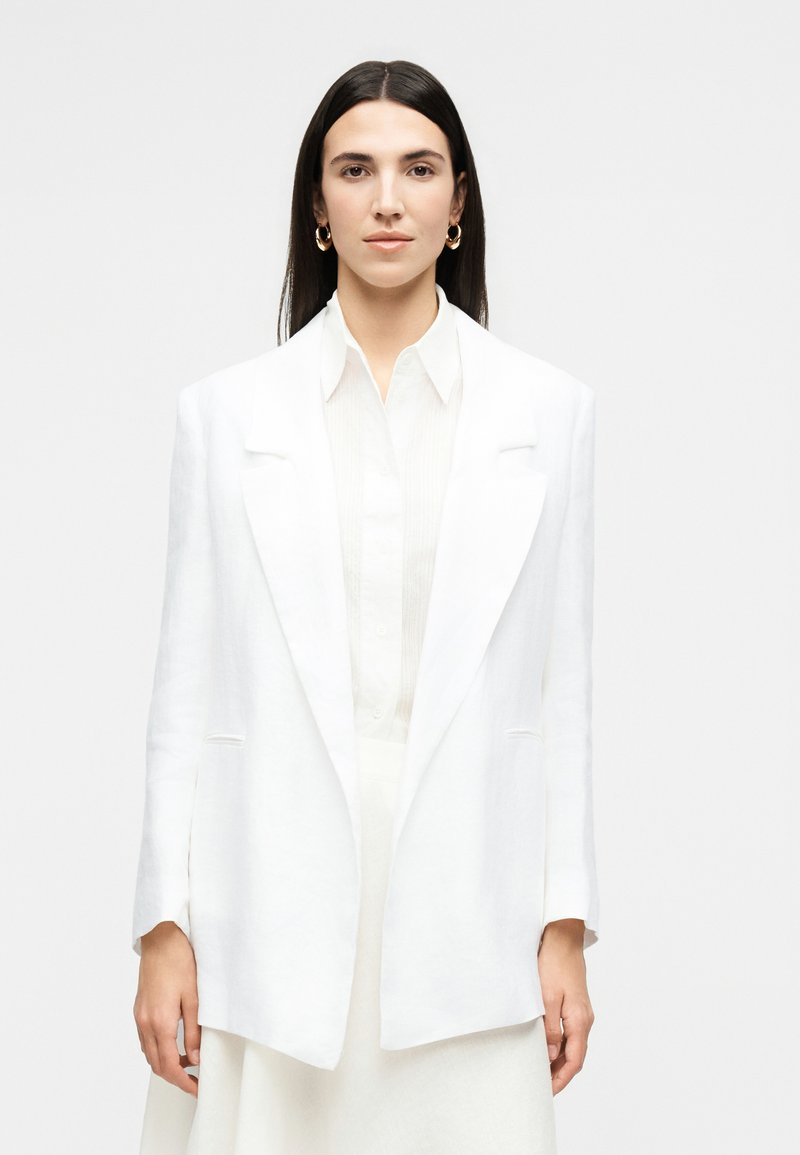 Γυναίκα με ίσια σκούρα μαλλιά που φοράει χρυσούς κρίκους σκουλαρίκια, λευκό πουκάμισο με γιακά και φαρδιά λευκή σακάκι, κοιτάζει μπροστά σε απλό φόντο.