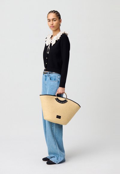 Bolso de paja tejido con un borde superior negro, asa circular y tachuelas decorativas, combinado con un suéter negro y jeans de mezclilla azul claro.