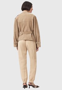 Frau von hinten, trägt eine hellbraune Wildlederjacke mit Gürtel, beige Hosen mit Gesäßtasche und Leopardenmuster-Pumps vor hellem Hintergrund.