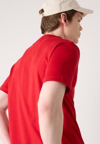 T-shirt en coton rouge à manches courtes, avec un col rond. Le modèle porte une casquette beige, affichant un design simple et décontracté.
