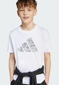 White cotton t-shirt featuring a gray camouflage Adidas logo in a geometric design. Crew neckline and short sleeves.