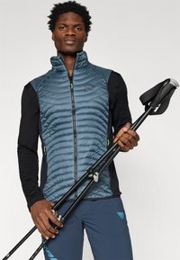 Μπλε μισοφόρι με καπιτονέ σχέδιο και μαύρους ώμους; συνδυασμένο με σκούρα παντελόνια και μπαστούνια πεζοπορίας κρατημένα σε μία χέρι.