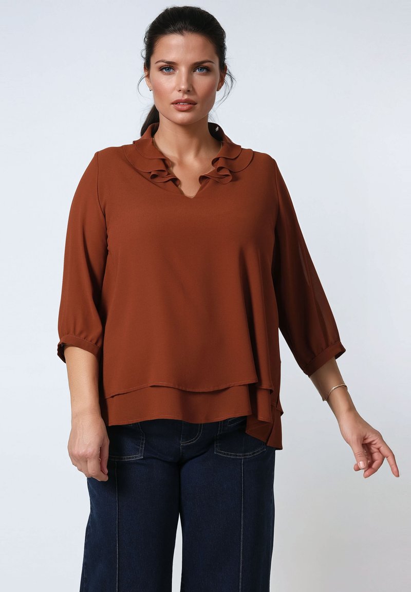 Vrouw met vastgebonden donker haar draagt een roestkleurige gelaagde blouse met een gerimpelde kraag en donkerblauwe jeans tegen een eenvoudige witte achtergrond.