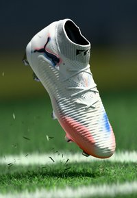 Biele futbalové kopačky s červenými a modrými akcentmi, čierne logo Puma, vo vzduchu nad trávnatým ihriskom vykopávajúce trávu.