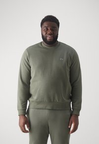 Felpa girocollo verde oliva con finitura texturizzata e polsini a coste, caratterizzata da un piccolo logo Lacoste sulla parte sinistra del petto.