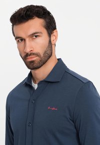 Marineblauwe lange mouw polo met een knoopkraag, voorzien van een klein rood geborduurd logo op de borst en een gladde, gestructureerde stof.