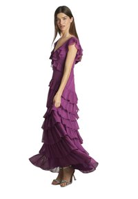 Lila, bodenlanges Kleid mit Rüschenschichten, kurzen Ärmeln und tailliertem Oberteil. Getragen mit silbernen, schnörkeligen High-Heels.