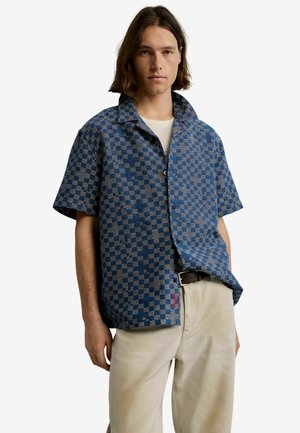 Młody mężczyzna w niebieską kratę, krótkim rękawie koszuli na białym t-shircie i beżowych spodniach, stojący na tle białego, gładkiego tła.