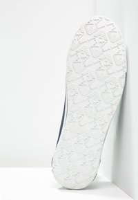 Weiße Gummisohle des Schuhs mit einem strukturierten Muster aus geometrischen Formen. Die Seite ist mit dunkelblauem Material versehen, das kontrastreiche weiße Akzente aufweist.