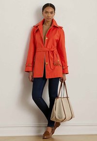 Ženska v oranžnem plašču s pasom, temnoplavih ozkih hlačah, rjavih loafers čevljih, drži bež torbo s rjavimi ročaji.