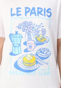 Valkoinen t-paita, jossa on sininen ja vaaleanpunainen graafinen kuvitus, joka esittää kahvipannua, kukkia, pannukakkuja ja croissanttia, tekstillä "LE PARIS BREAKFAST CLUB."