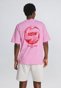 Pink bomulds t-shirt med et stort rødt grafisk motiv af en fodbold og teksten "HSW" samt "serve the game" på ryggen. Korte ærmer.