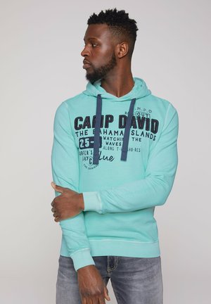 Sudadera verde menta con capucha, con cordones de marino y texto impreso. Tejido suave, puños y dobladillo acanalados, combinada con jeans de mezclilla.