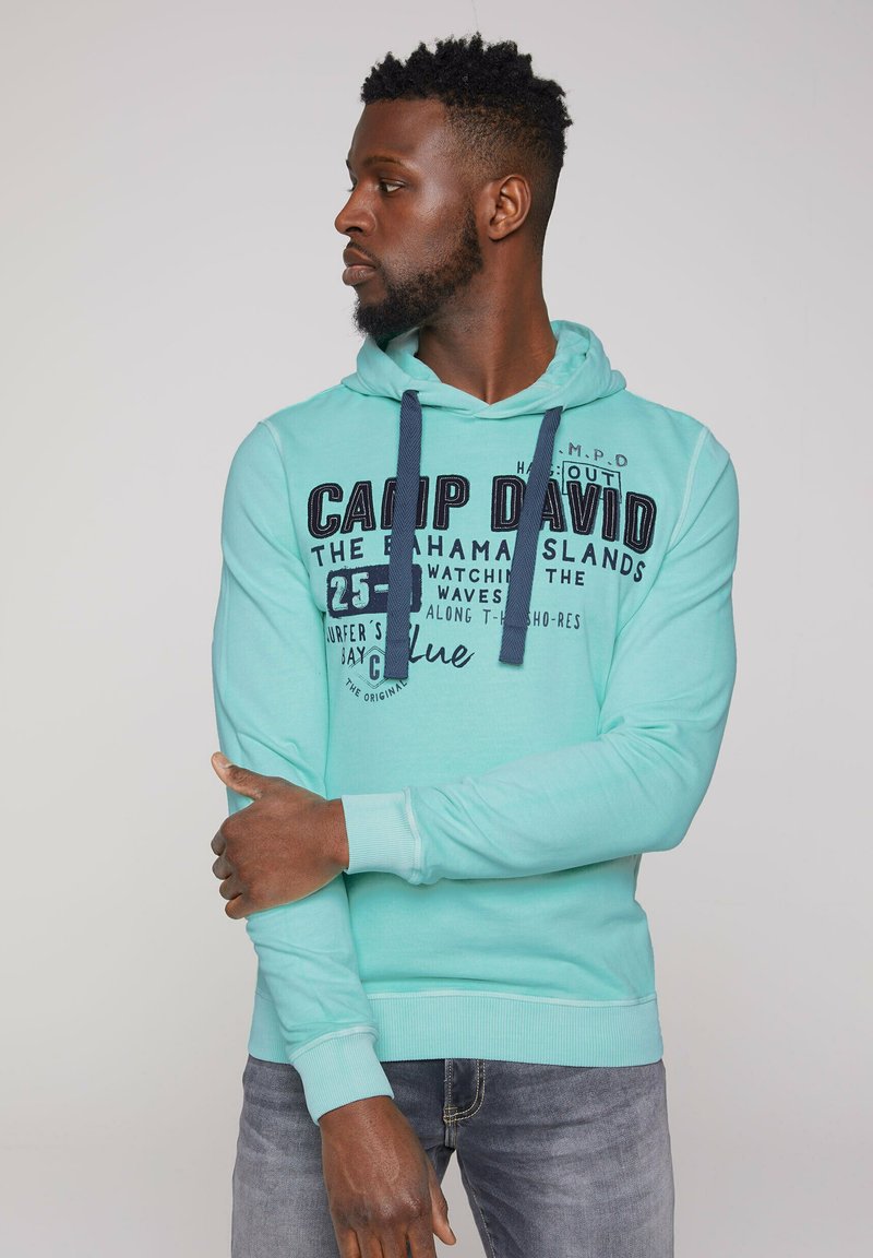Felpa verde menta con cappuccio, dotata di lacci blu navy e testo stampato. Tessuto morbido, polsini e orlo a coste, indossata con jeans in denim.