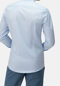 Chemise bleu clair à manches longues avec une texture lisse, un empiècement dorsal, des fentes latérales et un col classique. Portée avec un pantalon gris foncé.