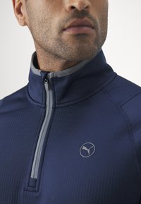 Pullover blu navy con colletto alto, zip grigia, texture a coste e logo discreto. Presenta un design aderente per l'abbigliamento sportivo.