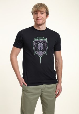 Junger Mann trägt ein schwarzes T-Shirt mit dem Text "Fierce Warrior" und einer Grafik eines kämpferischen Kriegers mit Helm sowie einer kleinen Kreatur darunter, kombiniert mit hellgrünen Hosen.