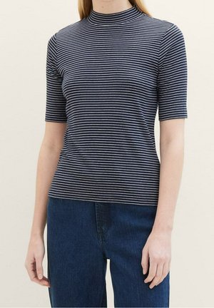 Kvinde iført en navy blå og hvid vandret stribet kortærmet bluse parret med mørkeblå denim jeans, stående mod en ensfarvet baggrund.