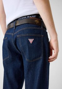 Osoba nosząca ciemnoniebieskie jeansy Guess z brązowym paskiem oraz białą koszulką na ramiączkach, pokazująca logo na tylnej kieszeni i metkę na pasku od spodni.