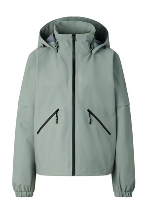 Junge Person trägt eine hellgraue Kapuzenjacke, rosa-grau gemusterte Leggings, schwarze Socken, schwarze Sneaker und eine schwarze rückwärts getragene Kappe und steht.