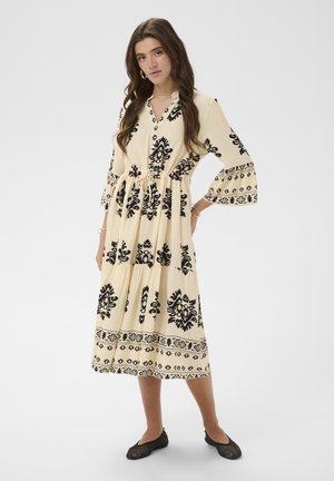 Vrouw met lang bruin haar, gekleed in een crèmekleurige midi-jurk met zwarte bloemenpatronen en zwarte gevlochten platte schoenen, staand met één hand op haar heup.