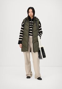 Gilet matelassé vert olive avec fermeture éclair et capuche, porté sur un haut à rayures noir et blanc. Un pantalon beige et un sac noir et crème complètent le look.