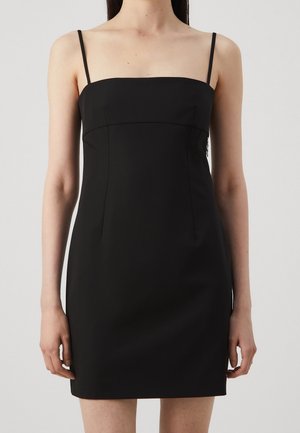 Mujer con un vestido mini negro sin mangas, con tirantes finos y escote recto sobre un fondo liso.