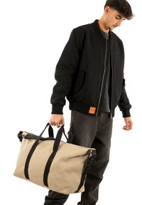 Borsa da viaggio impermeabile beige con cinghie nere, dotata di chiusura a rotolo e manici robusti. Il modello indossa una giacca bomber nera.