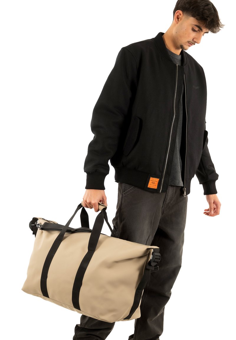 Borsa da viaggio impermeabile beige con cinghie nere, dotata di chiusura a rotolo e manici robusti. Il modello indossa una giacca bomber nera.
