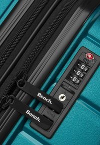 Türkisfarbene Hartschalenkoffer mit schwarzen Reißverschlüssen, einem Zahlenschloss und markierten Reißverschlussanhängern. Strukturierte Oberfläche mit faserigen Details.