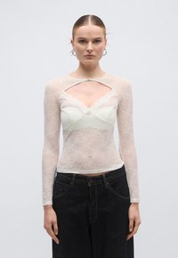 Μπλούζα με μακριά μανίκια από δαντέλα, με άνοιγμα σε σχήμα κλειδαρότρυπας, λευκό εσωτερικό στρώμα και περίπλοκο ανθισμένο μοτίβο. Συνδυάζεται με σκούρο τζιν παντελόνι φαρδιάς γραμμής.