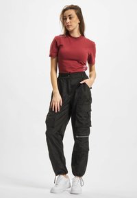 Bordeaux kortærmet skjorte parret med sorte cargo-bukser, der har lommer og lynlåse. Modellen har hvide sneakers på foran en ensfarvet baggrund.