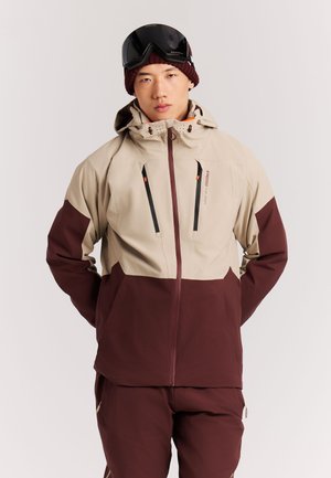 Junge Person trägt beige-braune Skijacke, passende Hose, dunkelrote Strickmütze und schwarze Skibrille auf der Stirn, steht mit Händen in den Taschen.