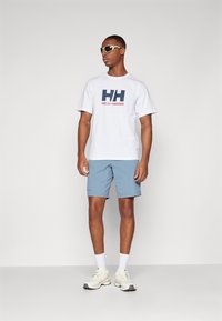 White T-shirt with navy and red "Helly Hansen" logo, paired with light blue shorts and white sports shoes. Model wearing sunglasses.