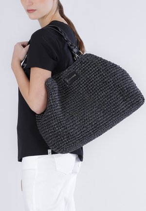 Mujer con camisa negra y pantalones blancos que lleva un gran bolso tejido negro con correas de cuero sobre el hombro.