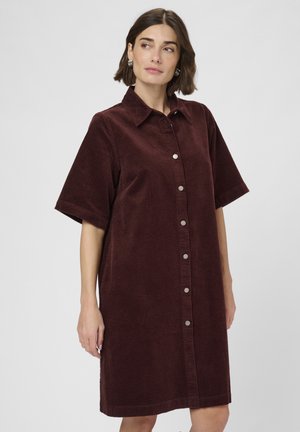 Femme aux cheveux bruns courts portant une robe chemise en velours côtelé bordeaux à manches courtes, se tenant devant un fond clair uni.