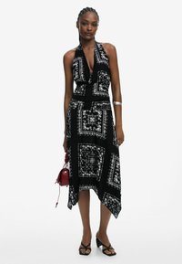 Schwarzes Halterkleid mit weißen Paisleymustern, asymmetrischem Saum und tailliertem Oberteil. Kombiniert mit einer kleinen roten Handtasche und schwarzen Sandalen.