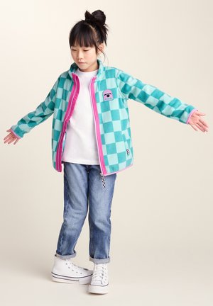 Child with hair in a bun wearing a turquoise checkered jacket, white shirt, cuffed jeans, and white high-top sneakers standing with arms outstretched.