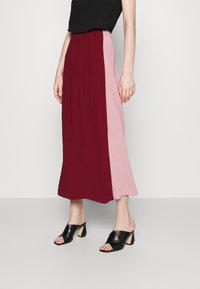 Kvinna klädd i en plisserad midikjol med burgundy- och rosa paneler, svart ärmlös topp och svarta klacksandaler mot en enkel bakgrund.