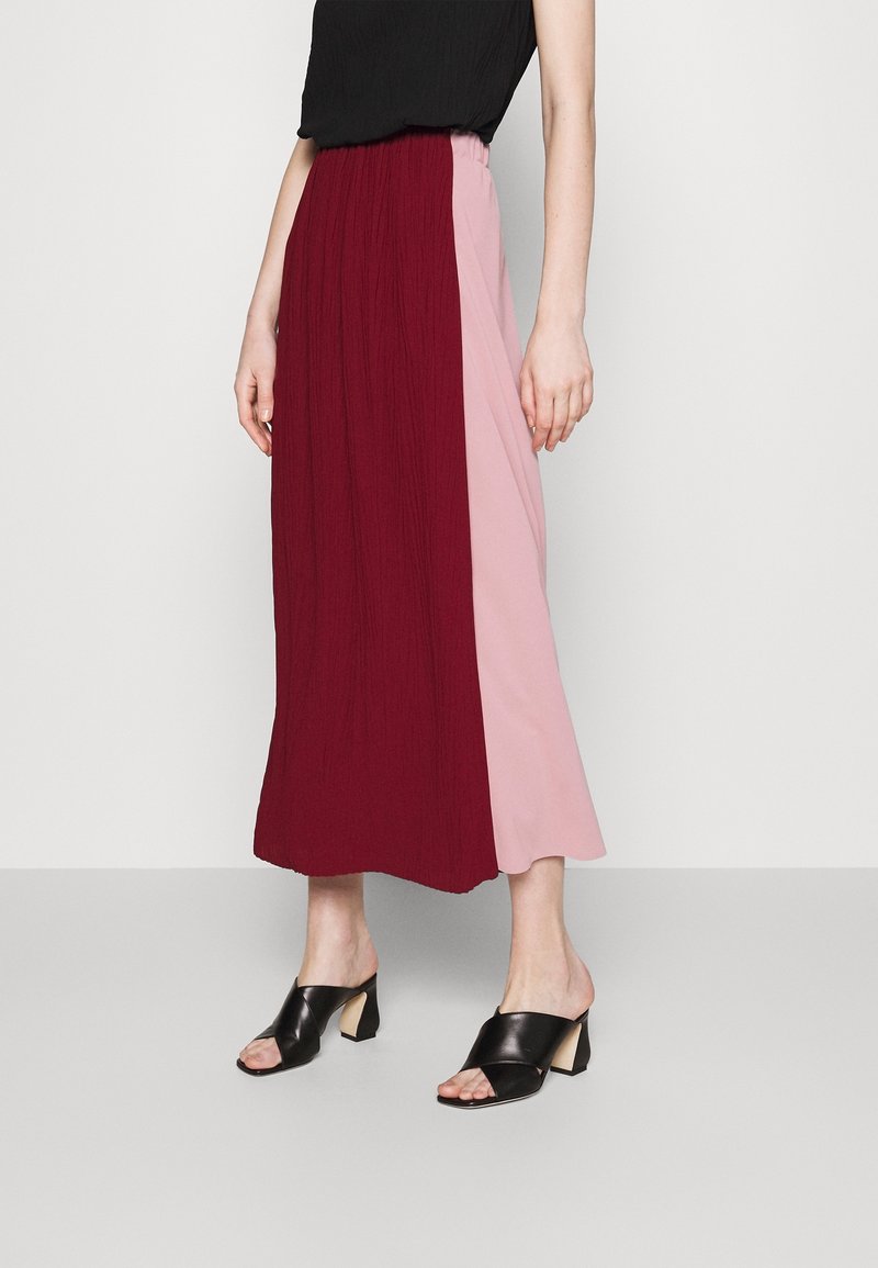 Kvinna klädd i en plisserad midikjol med burgundy- och rosa paneler, svart ärmlös topp och svarta klacksandaler mot en enkel bakgrund.