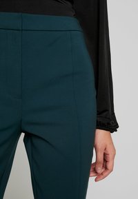 Turkosa högmidjade byxor i ett smidigt stretchtyg, med minimala sömdetaljer och en skräddarsydd passform. En hand vilar på midjebandet.