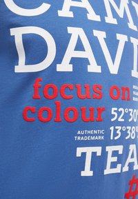 Blauer Stoff mit großem weißen und roten Text "DAVID focus on colour" und Koordinaten "52°30' 13°38'", Teil eines Grafikdesigns.