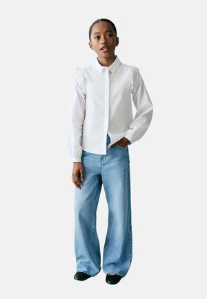 Chemise blanche à boutons avec des détails froncés aux épaules, portée avec un jean large bleu clair et des chaussures noires. Design simple et épuré.
