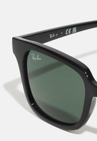 Slnečné okuliare Ray-Ban s štvorcovým čiernym plastovým rámom, zelenými zafarbenými šošovkami a logom Ray-Ban v ľavom hornom rohu šošovky.