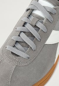Grå semsket sneakers med grå snørebånd, hvite detaljer og en gummirubel såle. Har en glatt tekstur og subtile stingdetaljer.